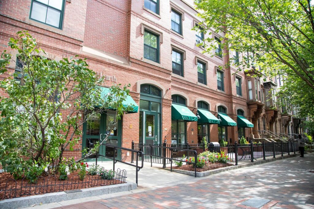 Older adult housing building with gardens out front shaded by trees, brick walls and green covered entries.