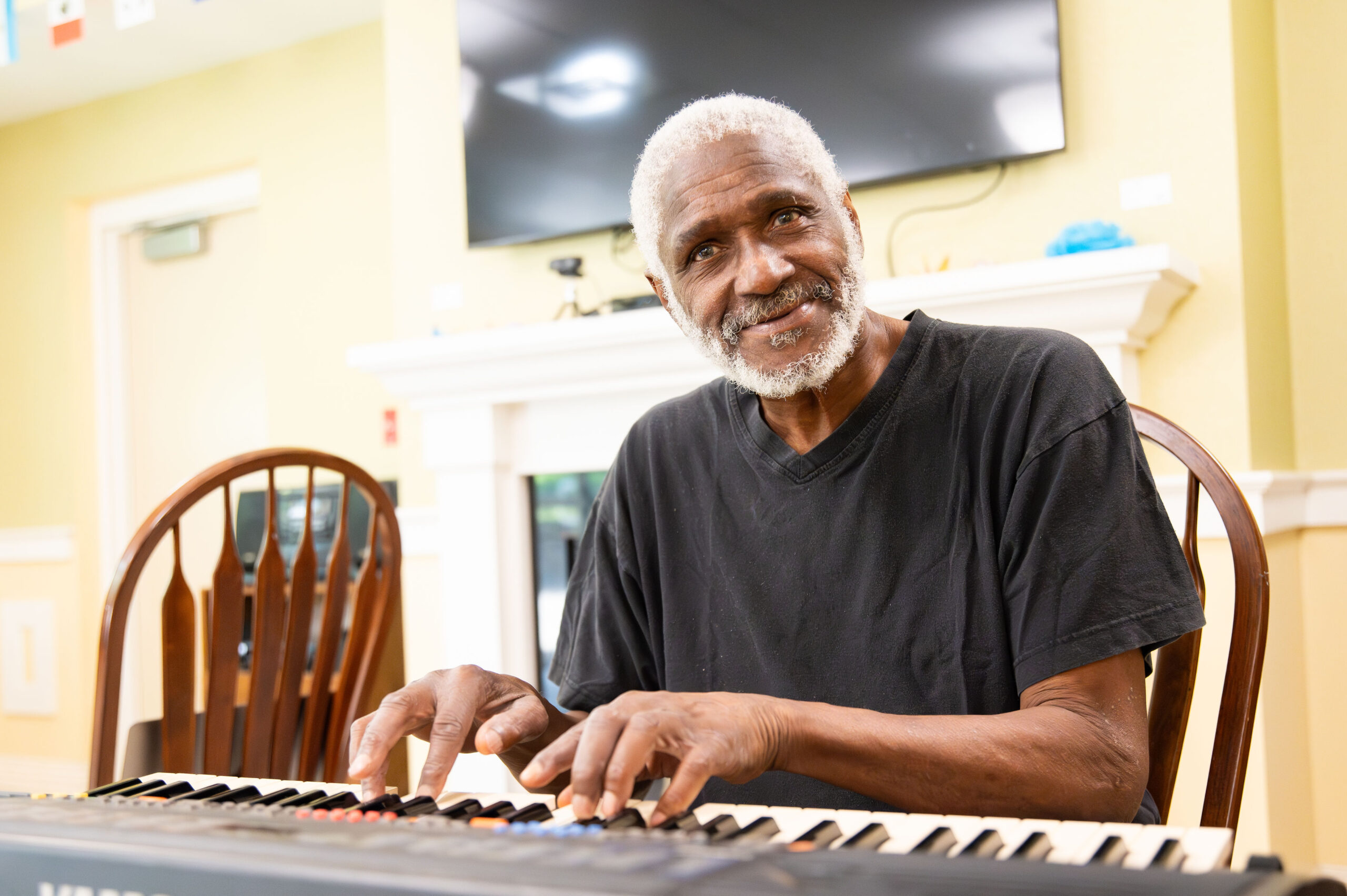 Older adult sitting at a table playing a keyboard looking at the camera and smiling.