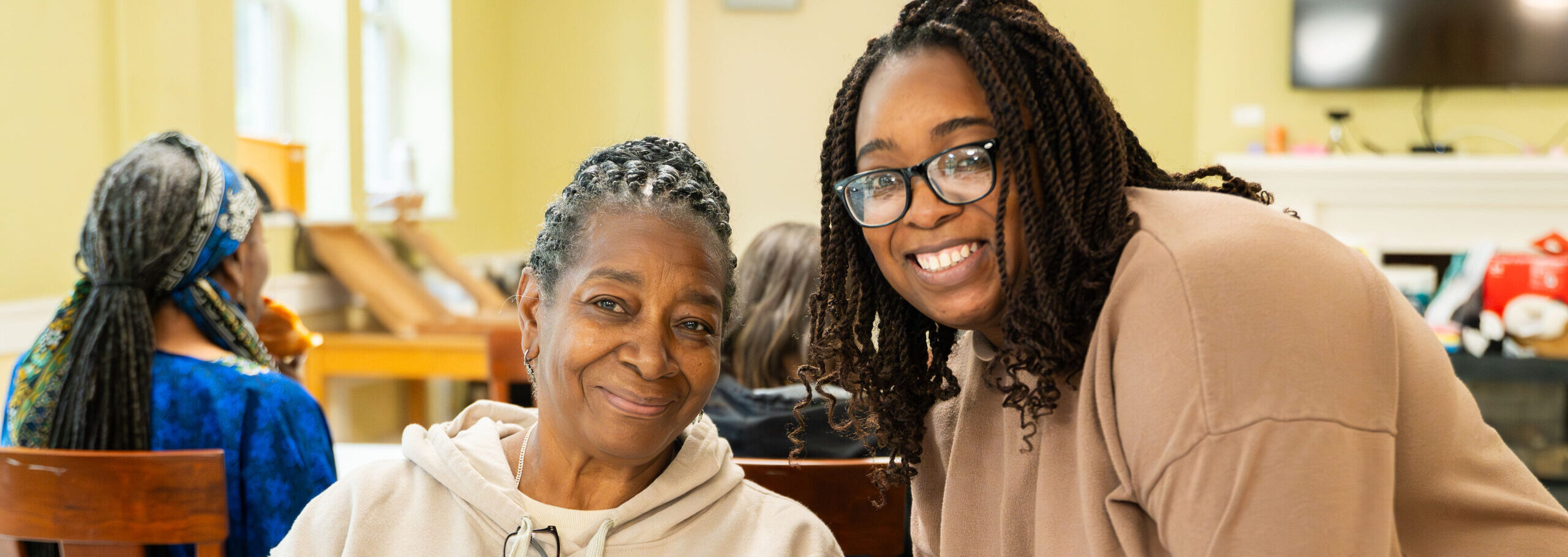 Two women, one older adult and one a younger volunteer, smiling and looking at the camera while seated in the living room of a Hearth Home Housing unit.